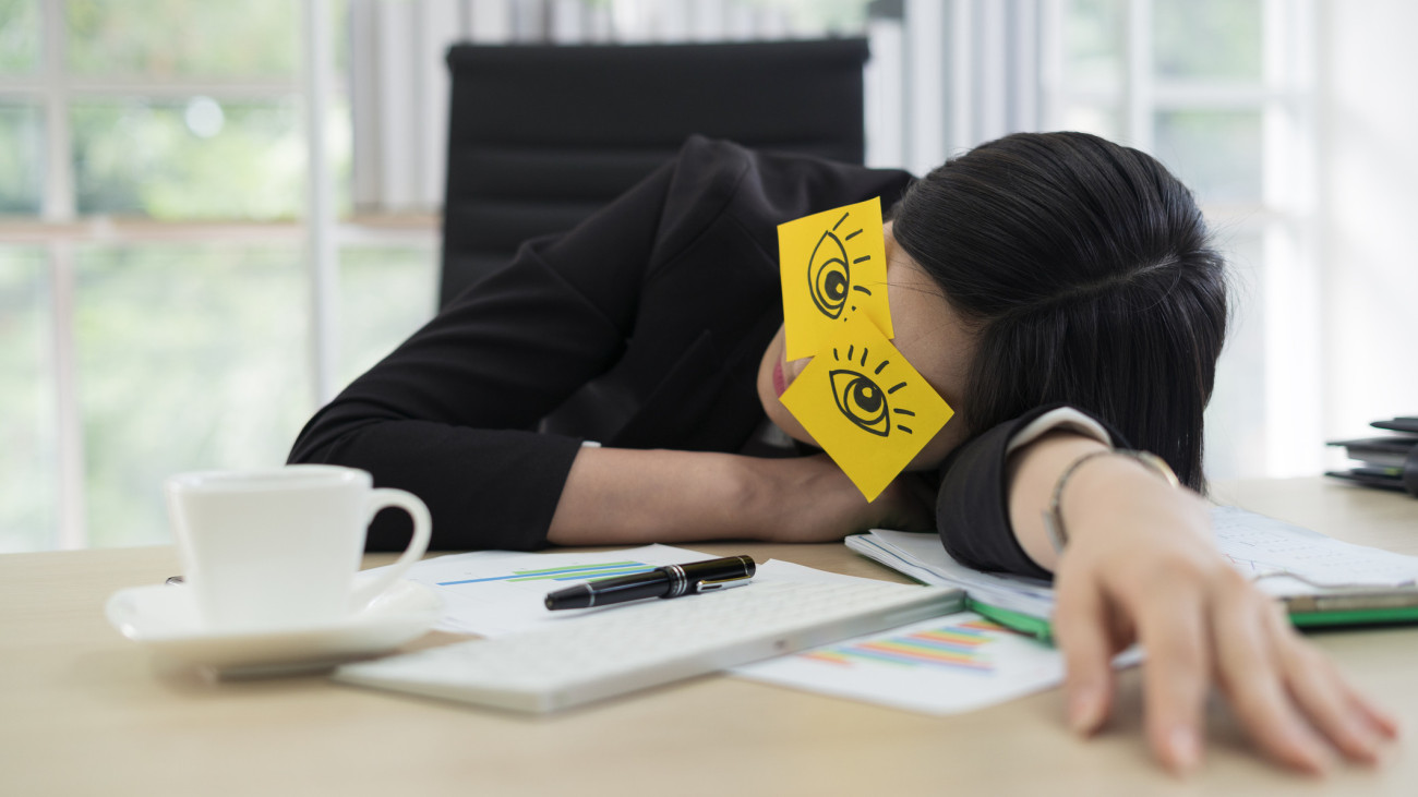 Young business woman with fake eyes painted on adhesive note sleeping at workplace in office