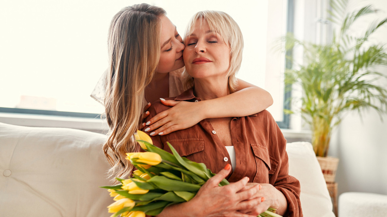 Happy Mothers Day and Womens Day. Charming daughter congratulating her mother, giving her a bouquet of tulips, hugging and kissing her in the living room by the window. Mothers love and care.
