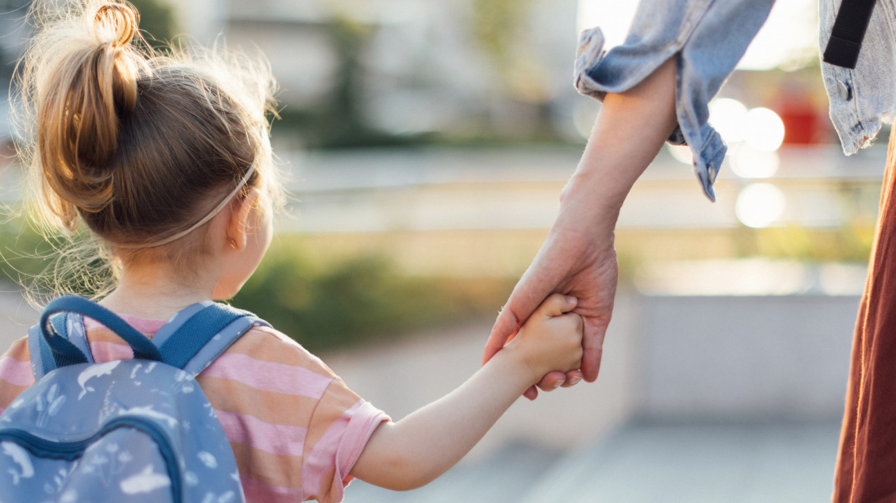 Back view of little girl with backpack holding a mom hand while walking to the school together.