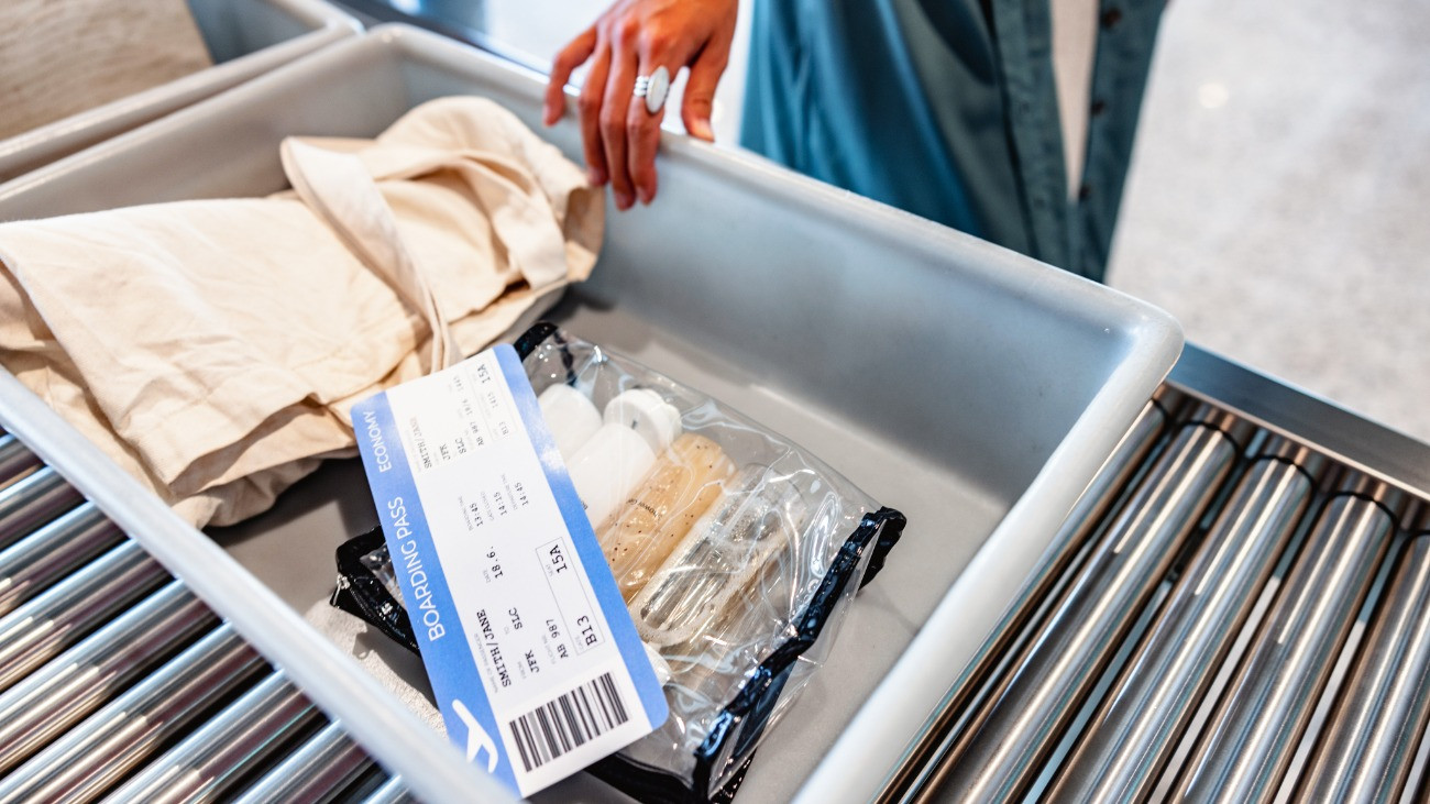 Biztonsági ellenőrzés egy repülőtéren, szürke műanyag tárolóban beszállókártya, folyadékok, textiltáska / Grey tray filled with liquid toiletries in a clear zip top bag, boarding pass and a beige tote bag. A female hand is holding the tray on a roller conveyors table at airport security.